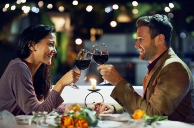 Smile, couple and cheers with wine at restaurant for date, love or holding hands together. Happy man, woman and toast with drink in glass for celebration, anniversary or romance with bokeh at night